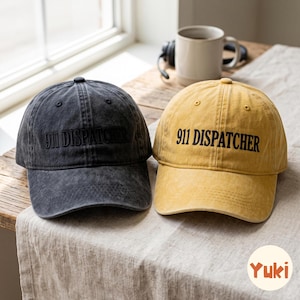May include: Two baseball caps, one in charcoal grey and one in mustard yellow, both featuring the embroidered text 911 DISPATCHER in black. Each cap has a structured crown, a curved brim, and a distressed, washed-out fabric finish.