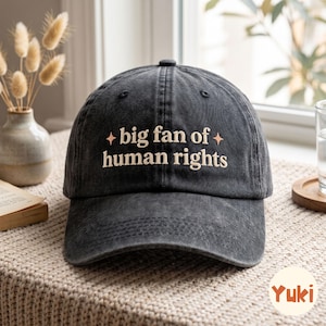 Może przedstawiać: Ciemnoszara czapka z daszkiem o wyblakłym, vintage wykończeniu. Z przodu znajduje się wyhaftowany jasnokremowy napis "big fan of human rights", otoczony po obu stronach dwiema małymi, czteroramiennymi ikonami gwiazdek.