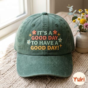 Può includere: Un cappellino da baseball verde bosco con il testo ricamato IT'S A GOOD DAY TO HAVE A GOOD DAY! Il design include piccoli fiori ricamati in rosa e azzurro, insieme ad accenti a forma di stella gialla, centrati sul pannello frontale.