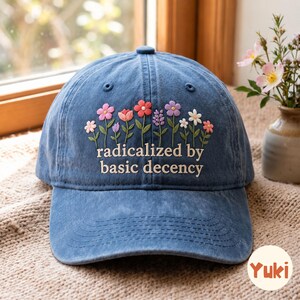 Könnte beinhalten: Eine blaue Baseballkappe mit gestickten bunten Wildblumen und dem Text "radicalized by basic decency" in weißer Schrift. Die Kappe hat eine strukturierte Krone, einen gebogenen Schirm und einen Knopf oben, was ein klassisches Sechs-Panel-Design zeigt.