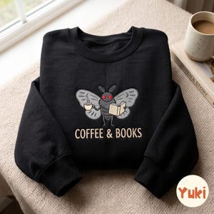 May include: A black crewneck sweatshirt featuring an embroidered graphic of a moth with red eyes, holding a steaming cup of coffee and an open book. Below the moth, the text COFFEE & BOOKS is embroidered in a light tan, sans-serif font.