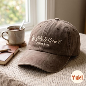 Könnte beinhalten: Eine braune Baseballkappe mit dem gestickten Text "Be Still & Know", gefolgt von "PSALM 46:10" und einem kleinen Herzsymbol. Die Kappe hat eine strukturierte Krone, einen gebogenen Schirm und sichtbare Nähte auf den vorderen Paneelen.