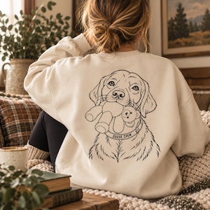 May include: A cream-colored crewneck sweatshirt featuring a black line art illustration on the back. The graphic depicts a dog holding a small teddy bear in its mouth, with a collar clearly displaying the text GOOSE CREW in capital letters.