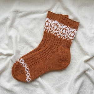 May include: A rust-coloured knit sock featuring a ribbed texture and a white geometric pattern around the cuff and toe. The sock is constructed with a reinforced heel and a standard crew length, showcasing a consistent colour palette throughout.