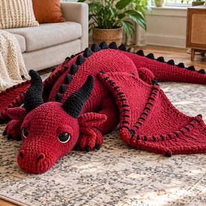 May include: A large, handmade crochet dragon plush toy in deep red yarn with black accents on the horns, wings, tail spikes, and heart-shaped tail tip. The creature features large, dark, glossy eyes and a textured, soft-looking yarn construction.