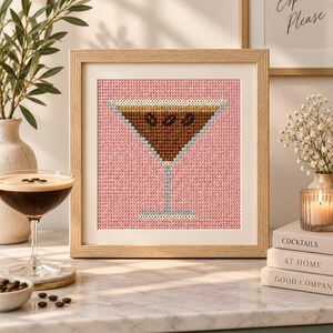 May include: A square framed cross-stitch artwork featuring a martini glass filled with a brown coffee-based cocktail, garnished with three dark brown coffee beans, set against a solid pink background within a light-coloured wooden frame.