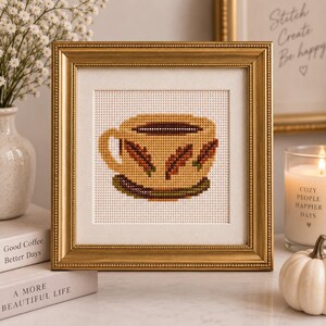 May include: A square cross-stitch embroidery featuring a tan coffee cup with brown leaf patterns, displayed in a gold-toned ornate frame. The cup sits on a dark green saucer, and the design is stitched onto a white fabric background.