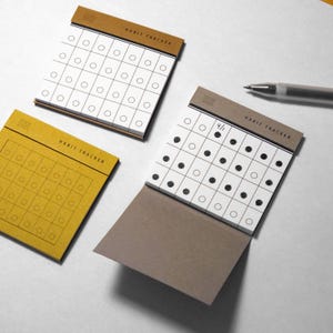 May include: Three square habit tracker notepads with a top binding. Two pads feature mustard yellow covers with a grid of circles, while one open pad has a grey cover and white pages with a grid of circles, some filled in with black ink.