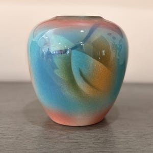 Puede incluir: Un pequeño jarrón de cerámica redondeado con un acabado liso y brillante con un diseño de degradado aerografiado. La superficie muestra colores mezclados de melocotón suave, turquesa vibrante, azul profundo y verde oliva en un patrón.