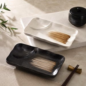Puede incluir: Dos platos rectangulares de cerámica con bordes redondeados, uno en color blanco roto y otro en negro, ambos con superficie texturizada y un patrón de pinceladas marrones. Cada plato incluye una pequeña hendidura circular en un lado.