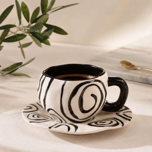 Pode incluir: Uma chávena de café em cerâmica com pires a condizer, apresentando uma forma irregular feita à mão, acabamento branco brilhante e padrões de espirais pretas pintadas à mão. A chávena possui uma asa preta espessa e arredondada.