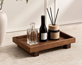 Könnte beinhalten: Ein rechteckiges Holztablett mit vier kurzen Füßen, auf dem sich eine Karaffe aus Klarglas, eine dunkle Glasflasche mit weißem Etikett und ein schwarzer Raumduft-Diffusor in einem dunklen Gefäß mit hellem Holzdeckel befinden.