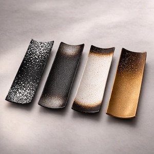 May include: Four rectangular ceramic chopstick rests with concave centers. The first is black with white speckles, the second is dark gray with a textured finish, the third is white with brown gradient edges, and the fourth is a metallic gold finish.