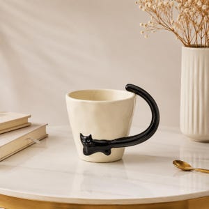 Può includere: Una tazza in ceramica color crema con una superficie irregolare e testurizzata. Un gatto nero lucido è applicato sul lato, con il corpo che forma la base del manico e la lunga coda curva che si estende verso l'alto per completarne la forma.