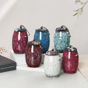 Può includere: Sei barattoli in ceramica con finiture smaltate lucide e crepate in tonalità rosso scuro, blu, turchese e bianco. Ogni barattolo presenta un corpo arrotondato e un coperchio rivestito in tessuto fissato con un cordino marrone e perline.