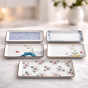 May include: A set of five rectangular ceramic plates with rounded corners and thin brown rims. The designs include blue wave patterns, blue floral borders, minimalist botanical illustrations with small red accents, and pink floral motifs.
