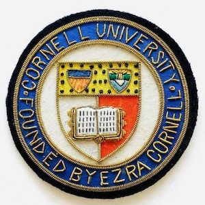 May include: A circular embroidered patch featuring the Cornell University seal. The design includes a shield with a book, a blue border with gold text reading CORNELL UNIVERSITY FOUNDED BY EZRA CORNELL, and a black outer edge.