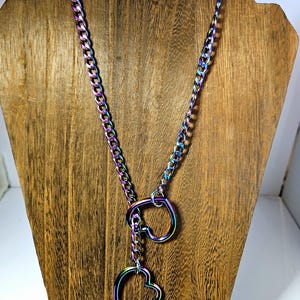 May include: A multi-colored chain necklace with two heart-shaped pendants. The necklace is displayed on a wooden jewelry stand. The chain and heart pendants have a rainbow iridescent finish, reflecting various colors.