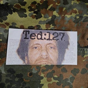 May include: A close-up of a printed paper with the text "Ted:127" in black and white. The text is surrounded by a blurry image of a person's face.