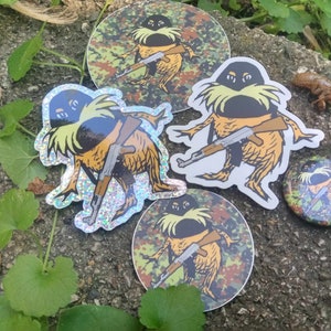 May include: Four colorful stickers featuring a cartoon character holding a rifle. The character has a green body, yellow clothing, and a black mustache. The stickers are on a gray and brown surface with green plants.
