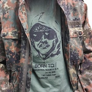 Può includere: Una maglietta verde con un'illustrazione in bianco e nero di una persona con occhiali da sole e una felpa con cappuccio. Il testo sulla maglietta recita "BORN TO INDUSTRIAL SOCIETY IS A Em All 1995 I am cabin man 54,235,754 CEOS".