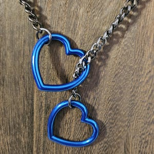 May include: A necklace featuring a silver chain and two bright blue heart-shaped pendants. The pendants are linked and hang from the chain. The background is a wooden surface, providing a contrast to the necklace's colours.