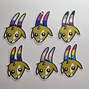 May include: A collection of six stickers featuring a cartoon goat with various pride flag horn designs. The goat has a tan body, white face, and black outline. The horns display non-binary, bisexual, asexual, rainbow, transgender, and pansexual pride flags.