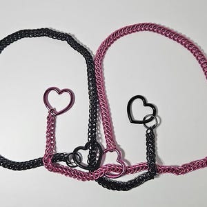 May include: Two chainmail necklaces with heart-shaped pendants. One necklace is black, the other is pink. The pendants are black and pink. The necklaces are connected with a chain.