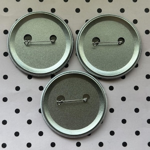 May include: Three circular metal pin-back badges arranged in a triangular formation. Each badge features a silver-toned metallic back with a horizontal pin mechanism secured through two small circular openings, showing the reverse side of the items.