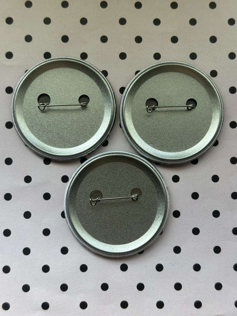 May include: Three circular metal pin-back badges arranged in a triangular formation. Each badge features a silver-toned metallic back with a horizontal pin mechanism secured through two small circular openings, showing the reverse side of the items.