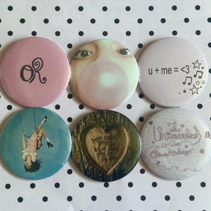 May include: A set of six circular pin-back buttons featuring various designs: a pink button with black 'OR' text, a face blowing a bubble, a white button with 'u + me = <3' and stars, a person on a swing, a heart in a tree, and 'The Unraveled' text.