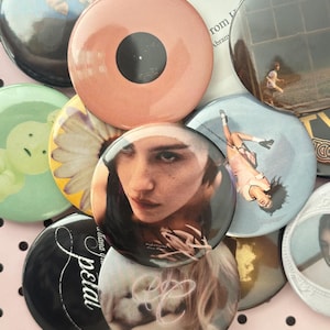 May include: A collection of circular pin-back buttons featuring diverse imagery, including a portrait of a person, a daisy, a puppy, a person running, and text such as 'petal' and 'from'. The buttons vary in color, including peach, black, white, and green.