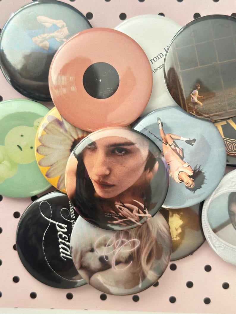 May include: A collection of circular pin-back badges featuring diverse imagery, including a portrait of a person, a daisy, a puppy, a person running, and text such as 'petal' and 'from'. The badges vary in colour, including peach, black, white, and green.