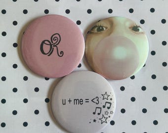 May include: Three circular pin-back badges. One is dusty pink with the black letters OR. Another features a photographic print of eyes and a bubblegum bubble. The third is white with the black text u + me = <3 and black star and music note icons.
