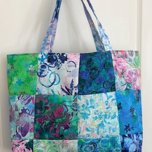 May include: A rectangular tote bag constructed from a patchwork of fabric squares featuring various floral and abstract patterns in shades of green, blue, teal, and pink. The bag includes two matching fabric shoulder straps attached at the top.