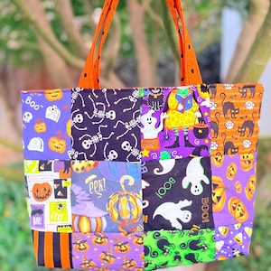 Può includere: Una borsa tote patchwork con vari quadrati di tessuto a tema Halloween in viola, arancione, nero e verde. I motivi includono fantasmi, scheletri, zucche, gatti neri e il testo "Boo" e "Eek". È dotata di due manici arancioni.