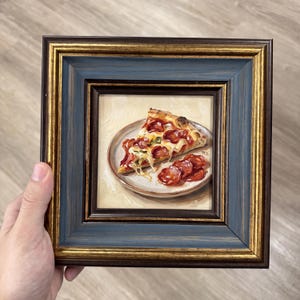 Könnte beinhalten: Ein quadratisch gerahmtes Ölgemälde, das ein Stück Pepperoni-Pizza auf einem Teller sowie zusätzliche Pepperoni-Scheiben zeigt. Der Rahmen ist dunkelblau mit Goldverzierungen und einer strukturierten Holzmaserung versehen.