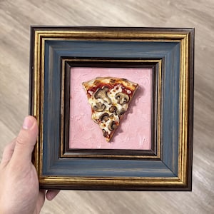 Könnte beinhalten: Ein gerahmtes dreidimensionales Kunstwerk mit einem Stück Pilzpizza, das auf einem strukturierten hellrosa Hintergrund zentriert ist. Der Rahmen ist dunkelbraun mit goldenen Innendetails und einem blaugrauen Innenrand.