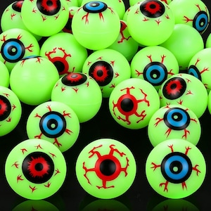Puede incluir: Una colección de pelotas saltarinas redondas de color verde lima con diseños de globos oculares impresos. Cada pelota muestra un iris central en rojo brillante o azul, rodeado por una pupila negra y patrones de venas rojas en la superficie.