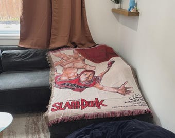 May include: A woven throw blanket featuring a graphic illustration of a basketball player in a red jersey reaching forward. The text Slam Dunk is printed in large red letters, with additional text including Takehiko Inoue and Weekly Shonen.