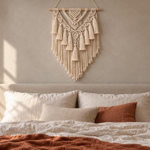 Puede incluir: Un tapiz de macramé de color crema con intrincados patrones de nudos, múltiples borlas colgantes y un borde inferior en forma de V. La pieza está suspendida de un listón de madera horizontal con un cordón blanco para su colocación.