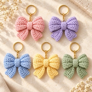 Puede incluir: Cinco llaveros de lazos de ganchillo en colores pastel: rosa, lavanda, azul claro, amarillo y verde salvia. Cada lazo está unido a una anilla de metal dorado mediante una cadena corta, con una textura de hilo tejido a mano.