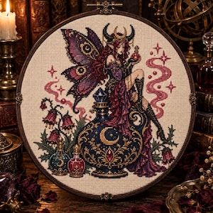 May include: A circular cross-stitch embroidery in a wooden hoop featuring a winged fairy with horns sitting on a large ornate blue potion bottle. The design includes purple butterfly wings, pink floral accents, and two smaller potion bottles.