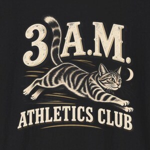 Könnte beinhalten: Ein schwarzes T-Shirt mit einem Grafikdruck einer laufenden getigerten Katze. Der Text 3 A.M. steht über der Katze und der Text ATHLETICS CLUB darunter. Das Design verwendet cremefarbene Schriftzüge und Katzendetails auf dunklem Grund.