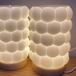 May include: Two cylindrical table lamps featuring a white, textured exterior composed of stacked, rounded, bubble-like shapes with fine vertical ridges. Each lamp sits on a circular base and includes a white power cord with an inline switch.