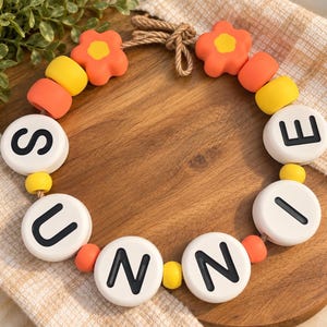 May include: A circular beaded garland featuring white round beads with the black letters S, U, N, N, I, E printed on them. The garland includes orange flower-shaped beads, yellow round beads, and smaller orange beads, all strung on a brown rope.