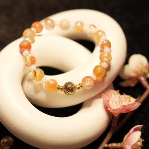 May include: A beaded bracelet featuring translucent orange and white agate stones interspersed with small clear beads and a central gold-toned metal bead with an etched floral pattern. The bracelet is circular and strung on an elastic cord.