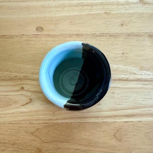May include: A small ceramic bowl featuring a two-tone design with a smooth light blue glaze on one side and a dark, glossy black glaze on the other. The interior of the bowl transitions between these two distinct colors, creating a sharp contrast.