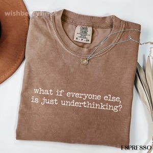 May include: A brown short-sleeve t-shirt with a crew neckline and a Comfort Colors brand label. The shirt features the white printed text: what if everyone else, is just underthinking? A silver chain necklace with a small circular pendant rests on the fabric.