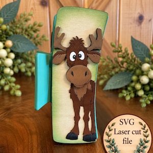 May include: A teal hair claw clip featuring a layered wooden moose cutout with brown fur, large antlers, and wide eyes. A circular wooden tag in the bottom right corner displays the text SVG Laser cut file surrounded by a green leaf border.